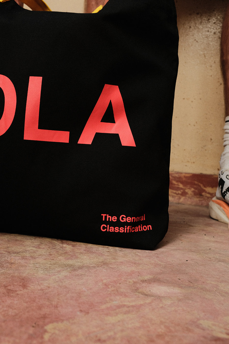 A black canvas tote bag with bold red text reading 'HOLA' and smaller red text 'The General Classification' near the bottom, photographed on a terracotta-colored floor against a textured wall. A partial foot/shoe is visible at the right edge.