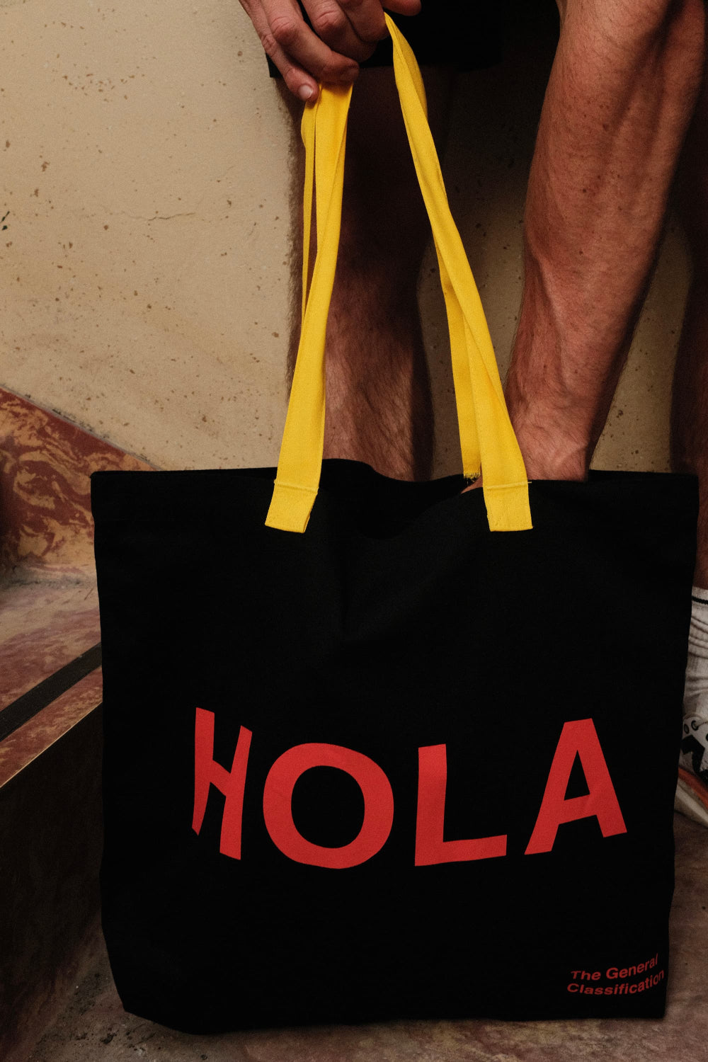 Black tote bag with red 'HOLA' text and yellow handles held by a person.