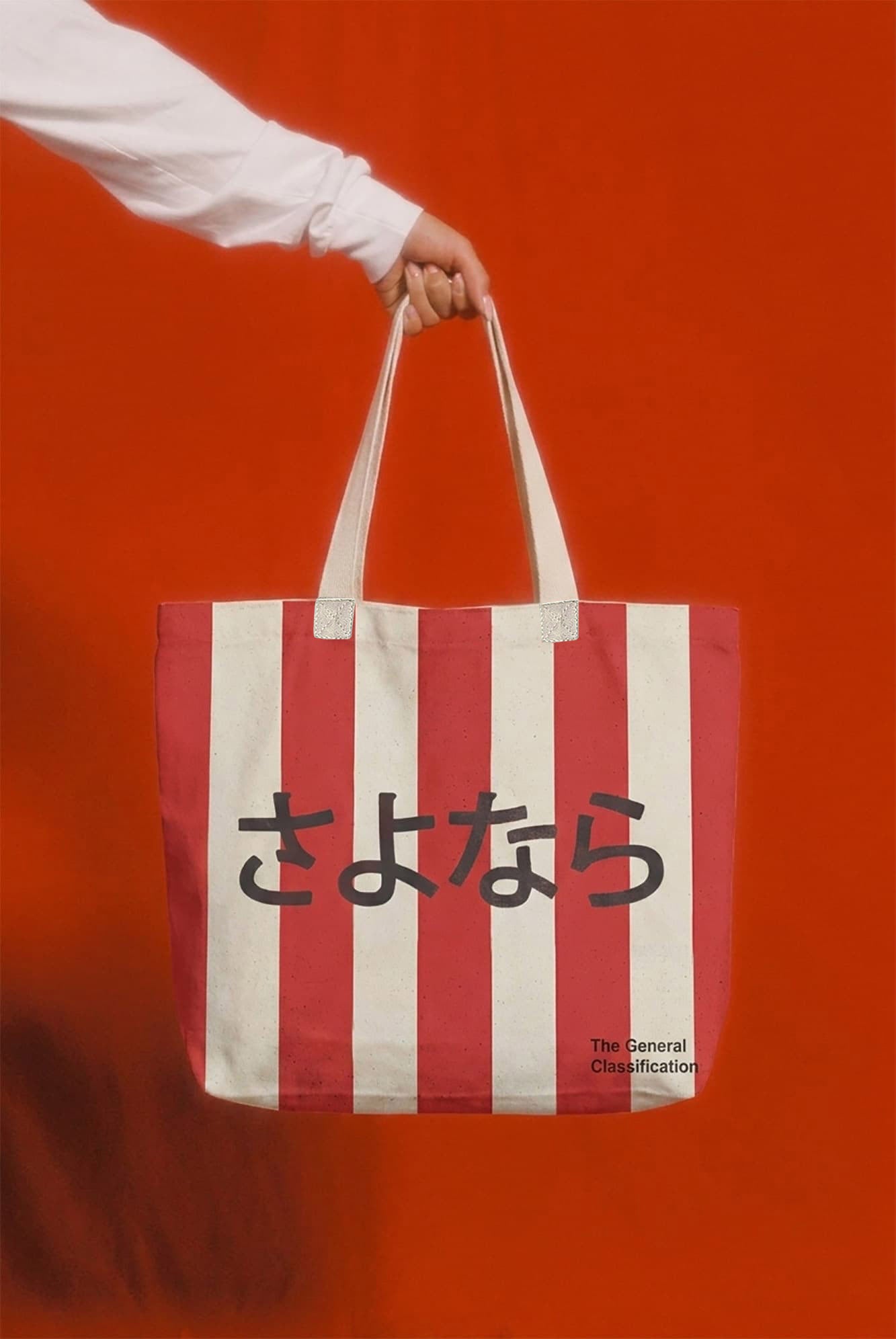 A striped tote bag with red and white vertical stripes, featuring the Japanese text konnichiwa in black across the center and 'The General Classification' printed in the bottom right corner. The bag is held by a person's hand wearing a white long-sleeve shirt, set against a solid orange background.