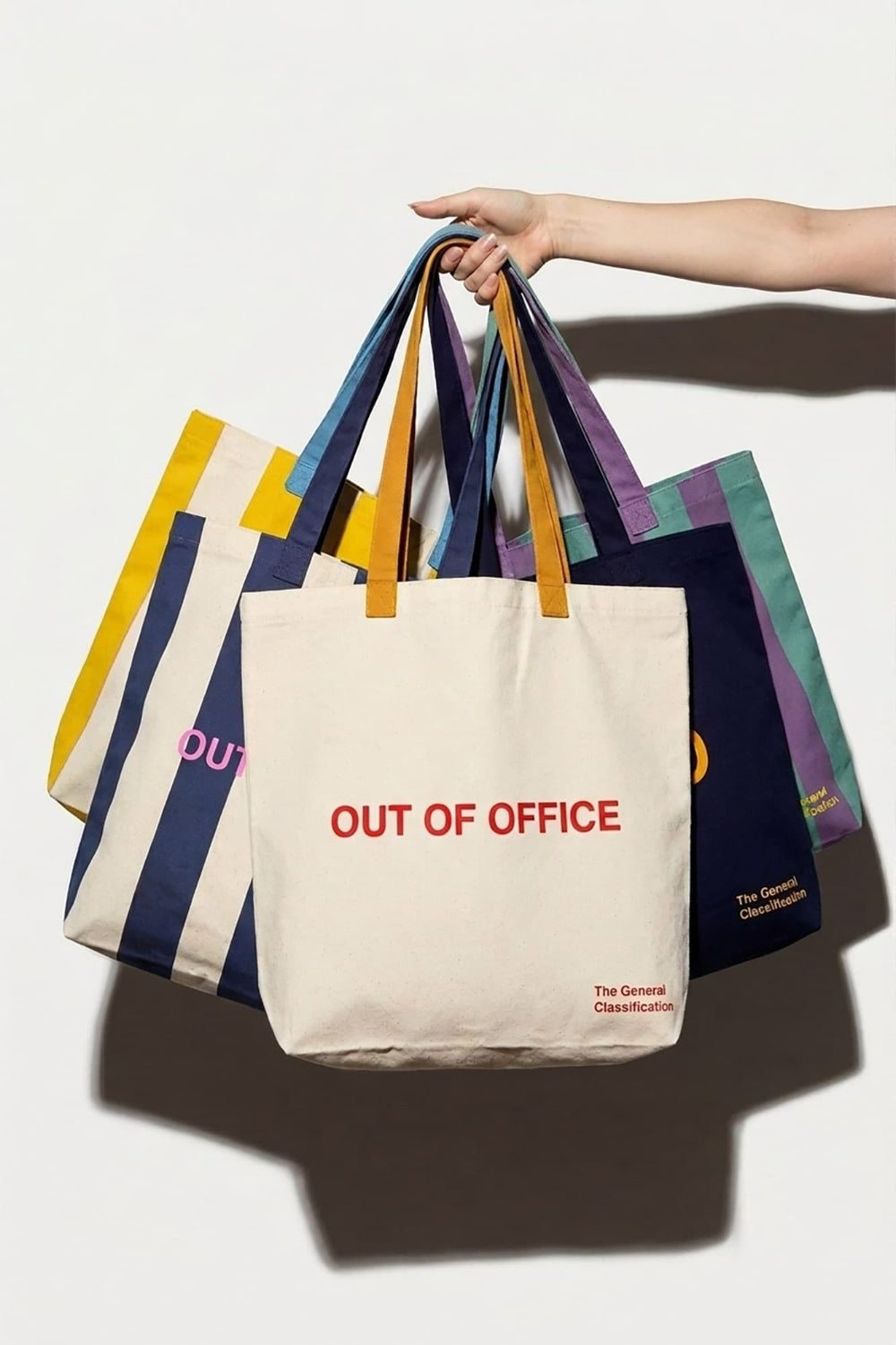 A hand holds a group of colorful canvas tote bags against a plain white background. The front tote bag is off-white with red text reading 'OUT OF OFFICE' and small red branding text at the lower right.