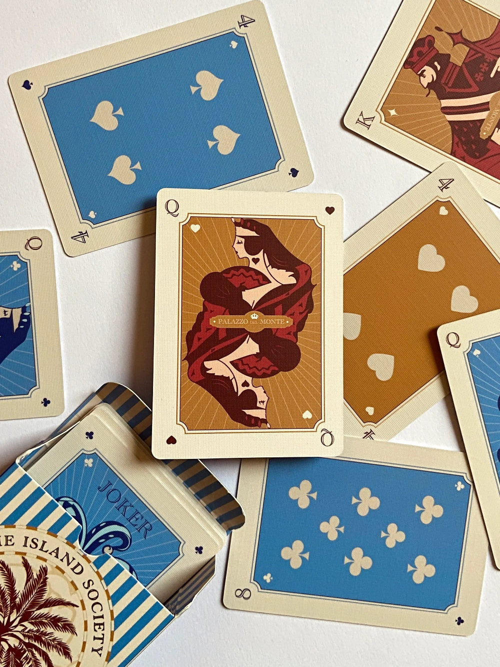 A styled flat-lay of vintage-style playing cards from The Island Society, featuring illustrated cards with cream borders, blue and gold backs, and a central Queen card showing a woman in a red dress.