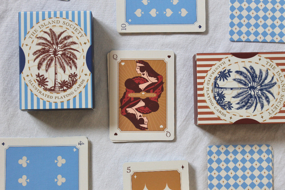 The Island Society illustrated playing cards arranged on a light fabric surface, showing card backs and a few face-up cards including a Queen of Hearts.