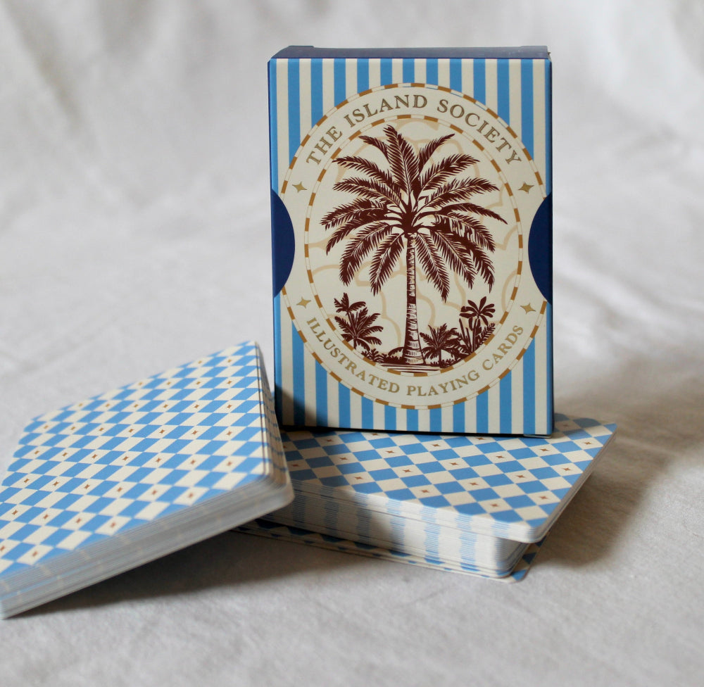 A boxed deck of illustrated playing cards standing upright with two smaller stacks in front. The box has blue and white vertical stripes and a cream oval label with a palm tree illustration and the text 'The Island Society Illustrated Playing Cards.
