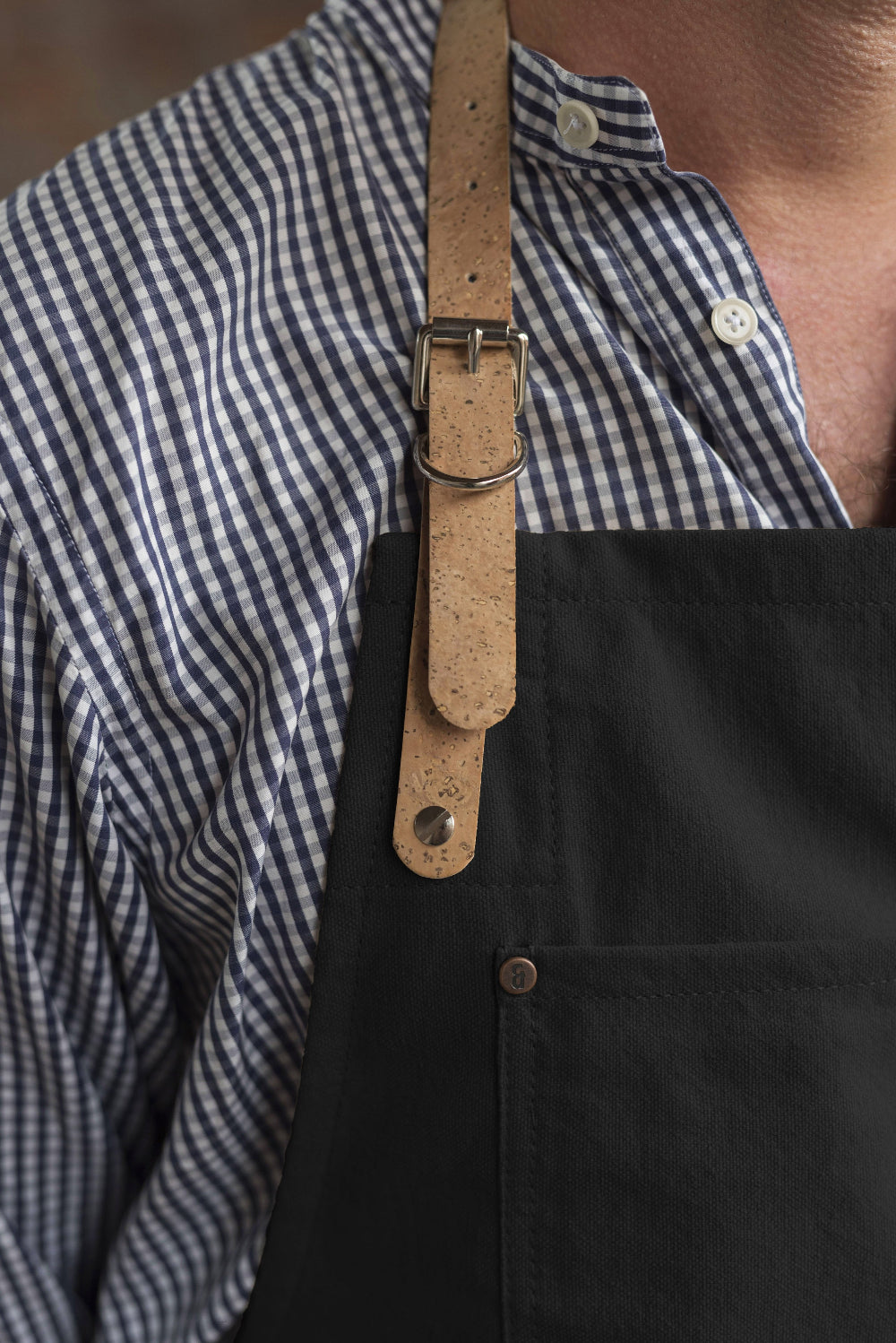 Person wearing a black apron with a brown cork strap over a checkered shirt.