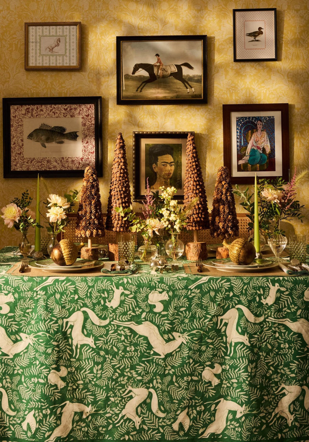 Decorative table setting with a green patterned tablecloth, candles, and framed pictures on a wall.