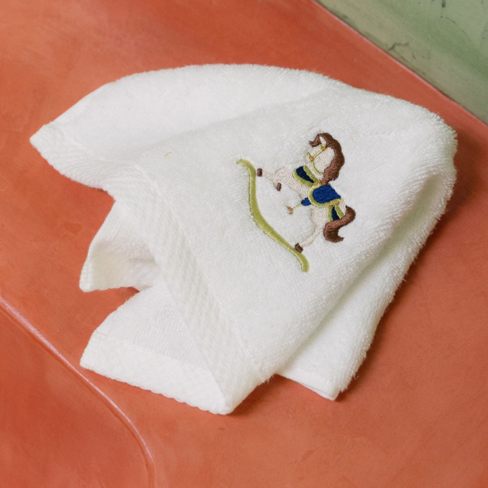 White towel with embroidered rocking horse green and blue design on a red surface