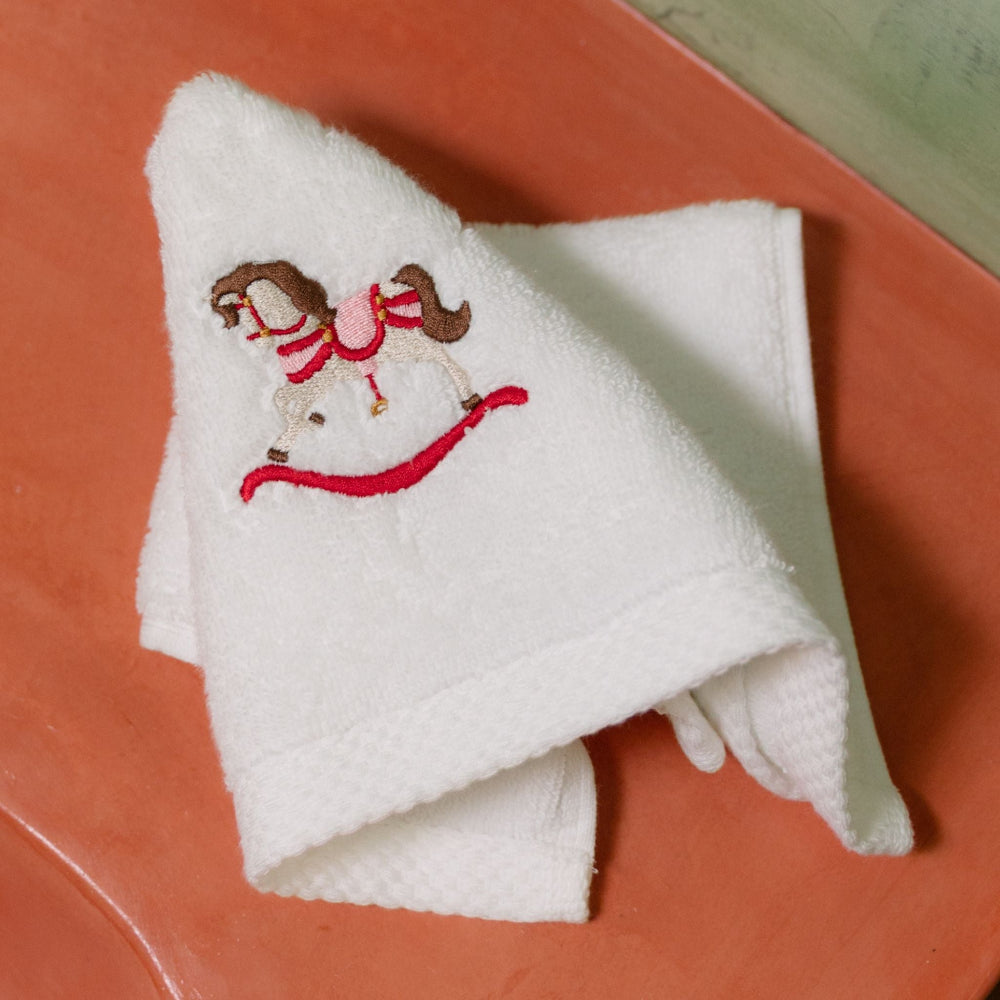 Folded white towel with embroidered christmas rocking horse design on a red surface