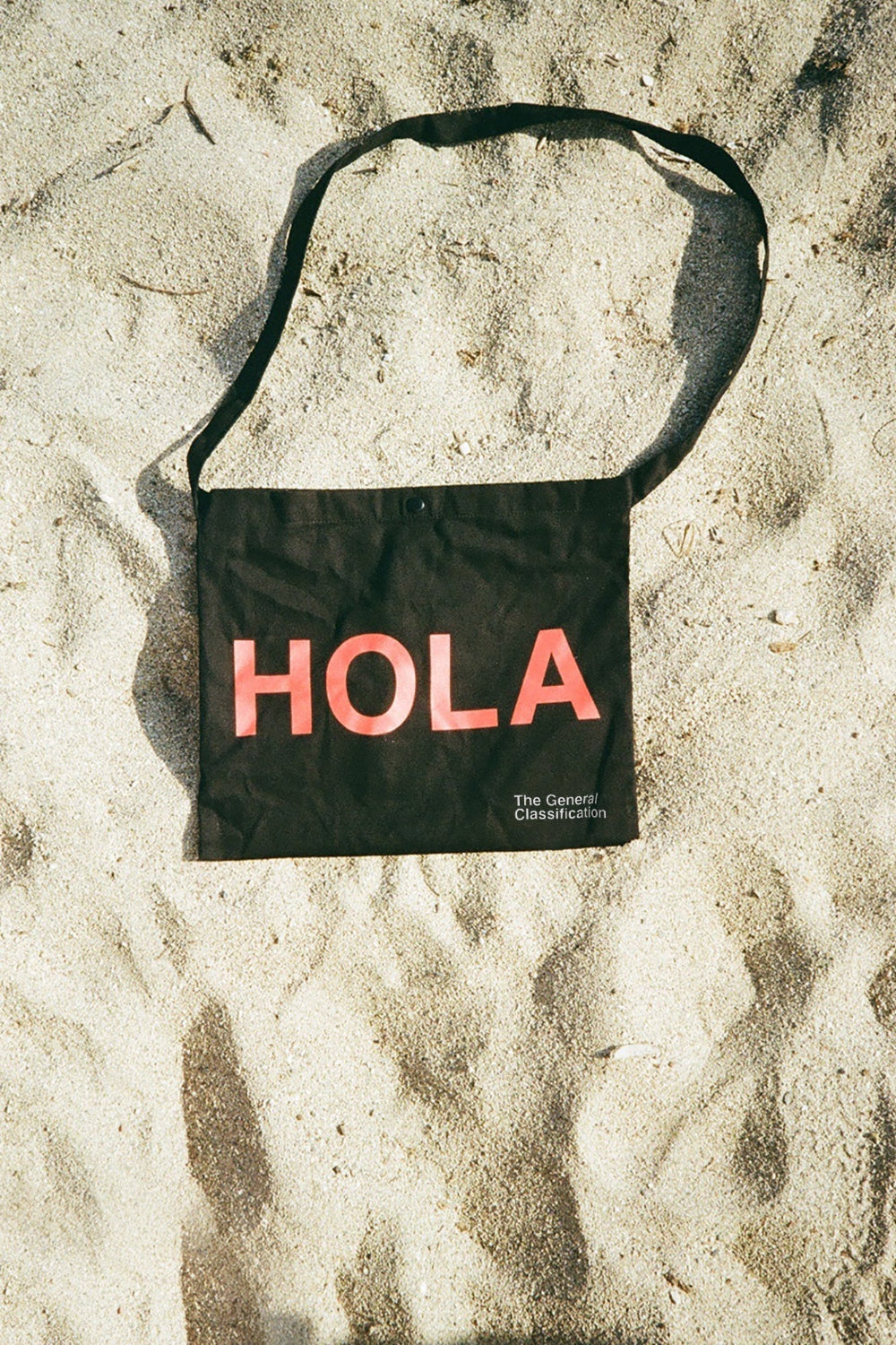 A black canvas shoulder bag with a long strap lies on sandy ground. The bag has the word 'HOLA' printed in large coral-pink letters, with smaller white text reading 'The General Classification' underneath.