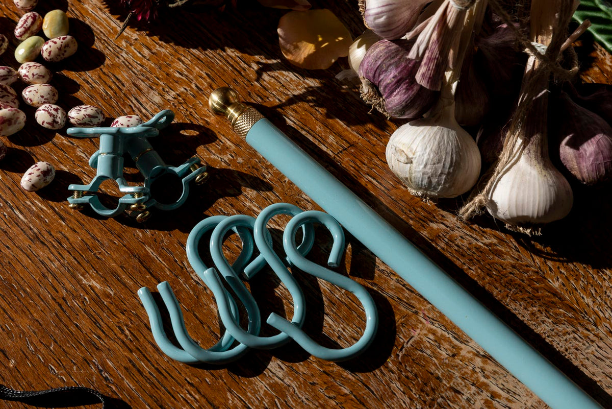 A light blue utensil rail with a brass finial and matching blue curtain rings, displayed on a rustic wooden table alongside garlic, quail eggs, and dried flowers.