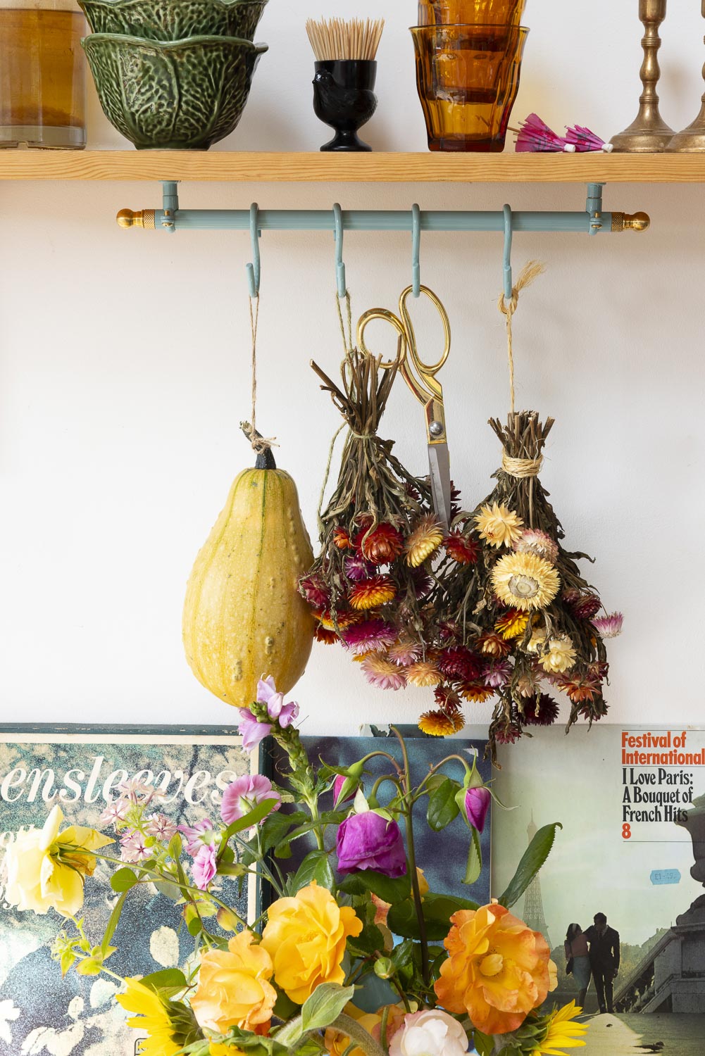 A rustic wall-mounted horizontal bar with hanging hooks holds dried botanical bundles, including a yellow squash and scissors, above a bouquet of yellow, orange, and purple flowers. A wooden shelf with glassware and decorative objects sits above against a white wall.