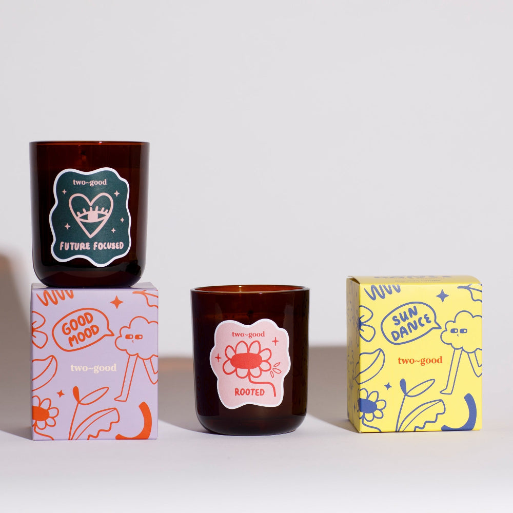 Two brown candles with decorative labels on a white background, surrounded by their packaging boxes.