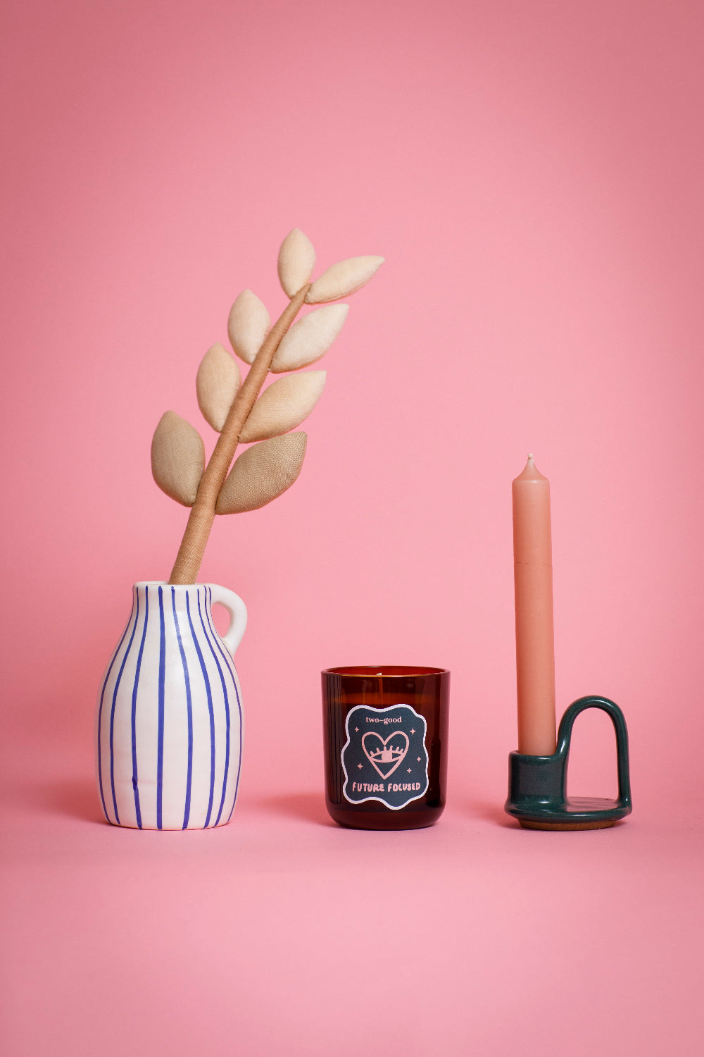 Decorative items including a striped vase, candle from two good, and a plant on a pink background