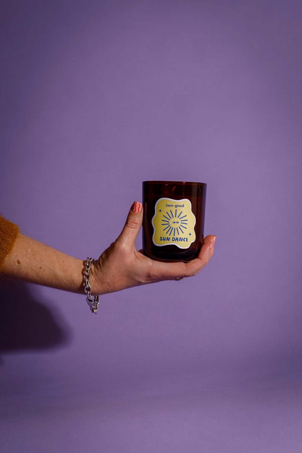 Hand holding a dark glass candle jar with a label 'Sun Dance' against a purple background