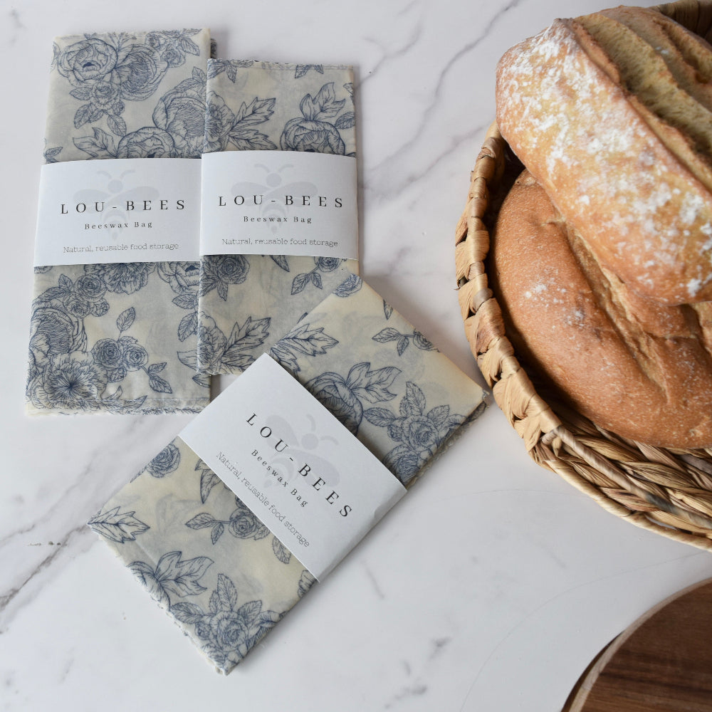 Floral-patterned beeswax bags branded Lou-Bees bags on a marble surface with bread in a basket.