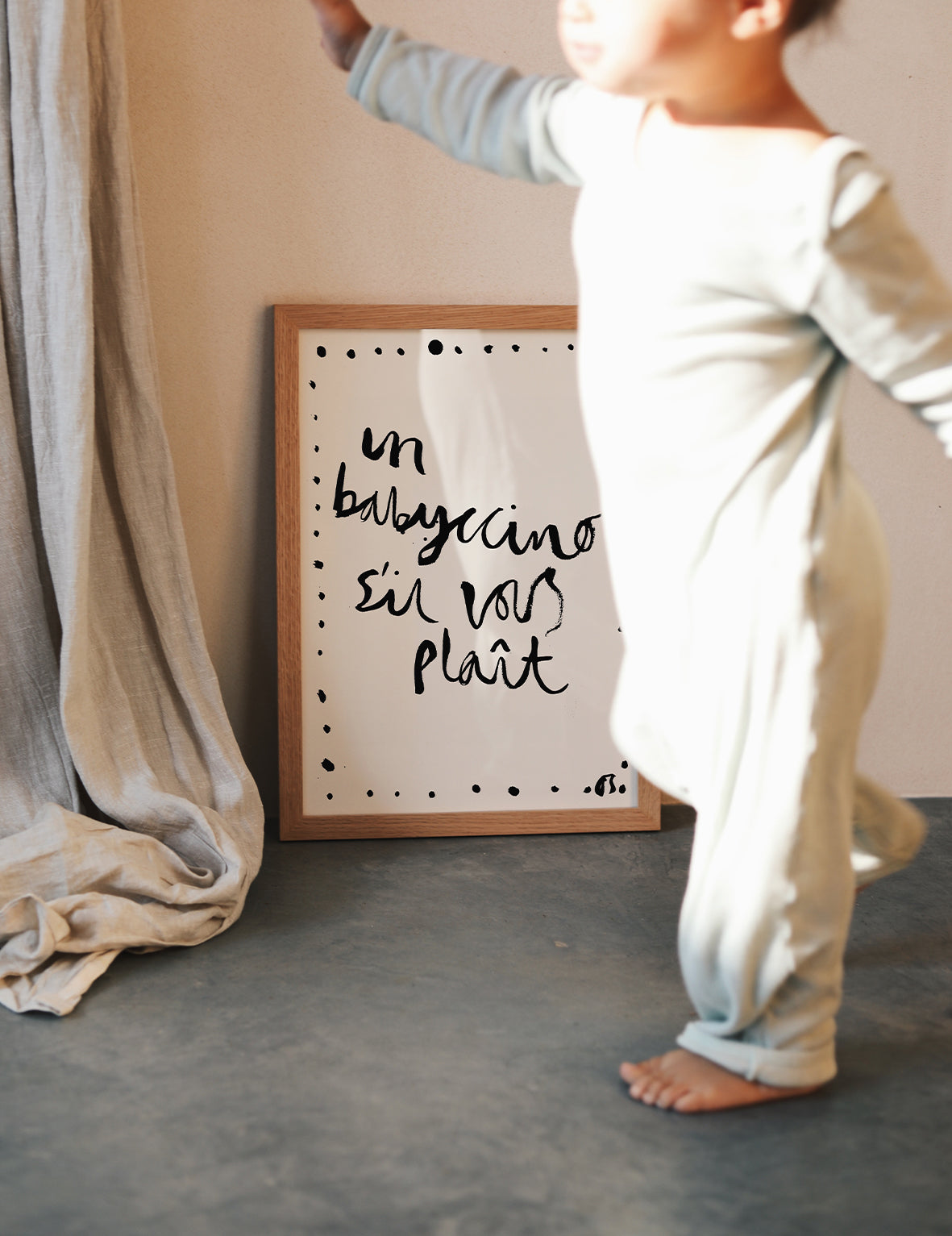 Child standing next to a framed poster 'un babyccino sil vous plait' text on a neutral background