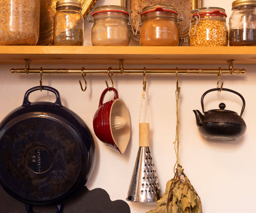 A brass kitchen wall rack with hooks hanging cookware and utensils beneath a wooden shelf filled with glass jars of spices and grains. Visible items include a dark blue enameled cast iron pot, a red ceramic mug, a metal cone grater, a black teapot, and a dried herb bundle.
