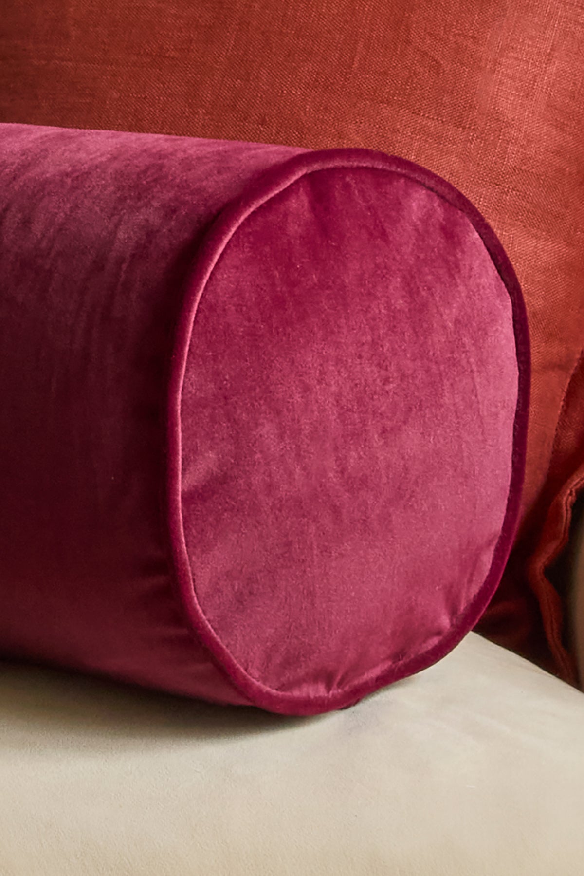 A close-up of a cylindrical lumbar pillow in deep magenta velvet, resting on a light beige surface against a rust-red fabric background.