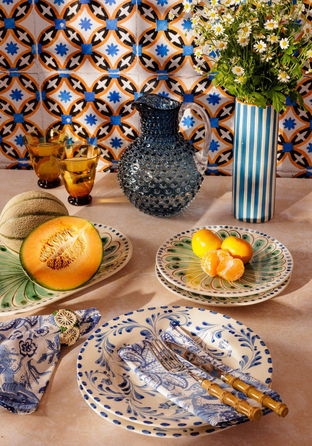 Dining table setting with blue and white ceramic plates, cutlery, and a decorative vase against a patterned tile wall.