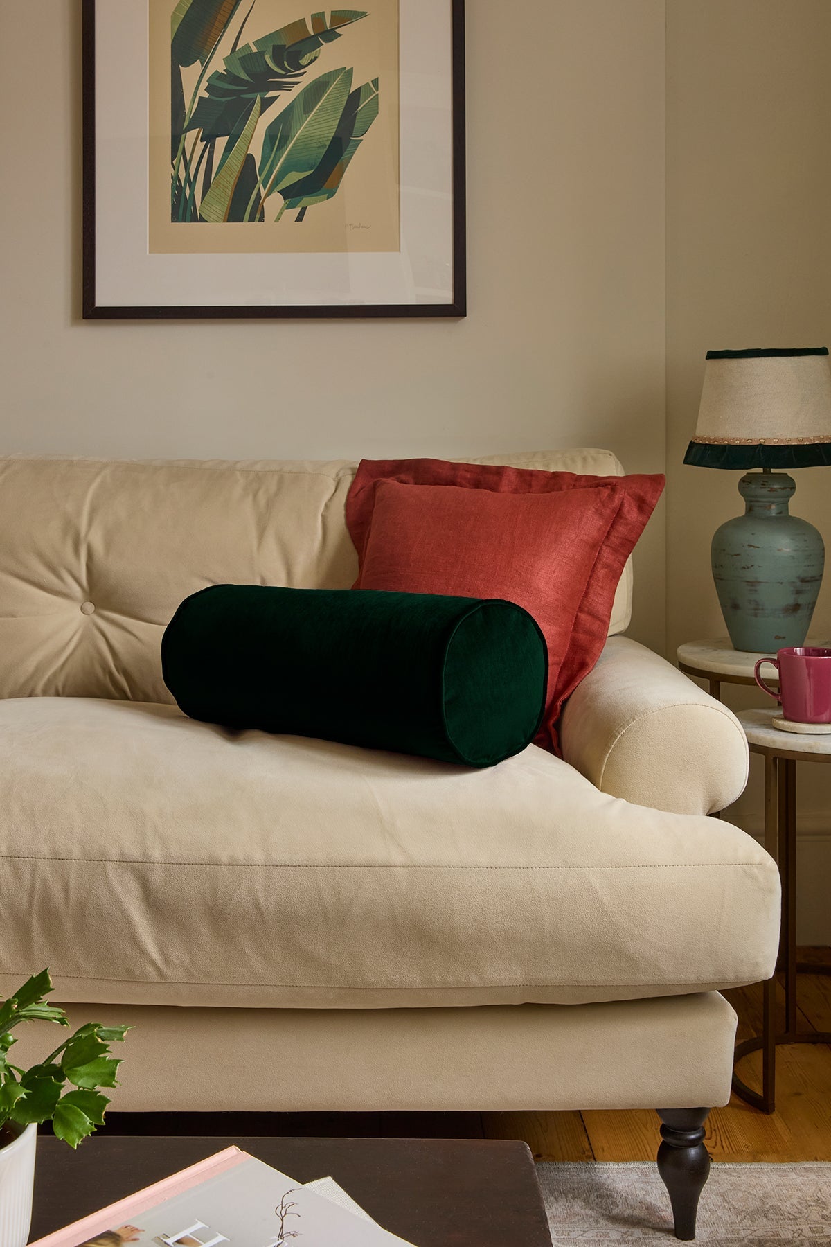 A styled living room scene featuring a cream tufted sofa with decorative pillows, including a dark green cylindrical lumbar pillow and a rust-red square throw pillow. A matching framed botanical print hangs on the beige wall above the sofa, and a blue-green ceramic table lamp sits on a side table next to a pink mug.