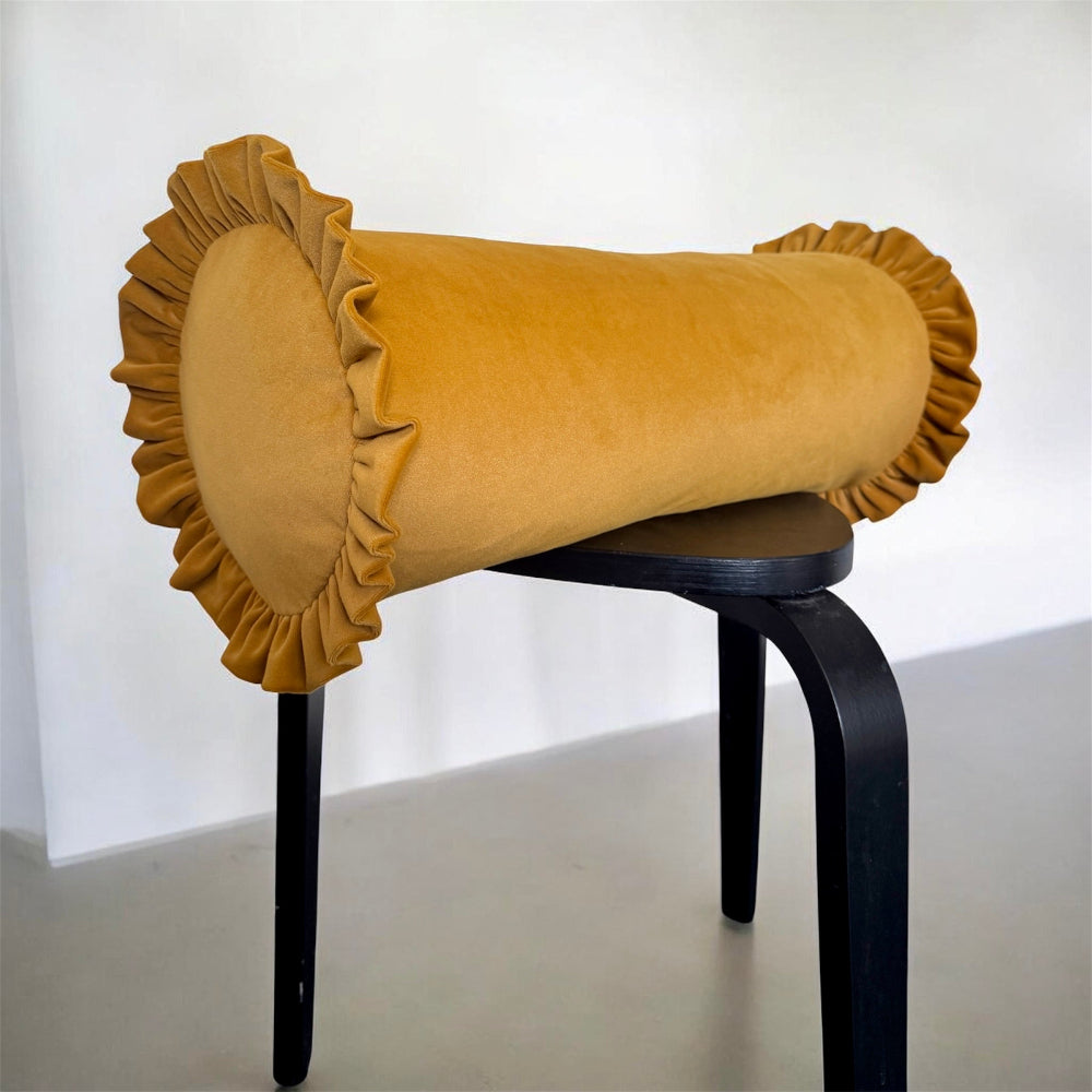 A mustard yellow velvet cylindrical pillow with ruffled trim, displayed on a black stool against a plain white wall.