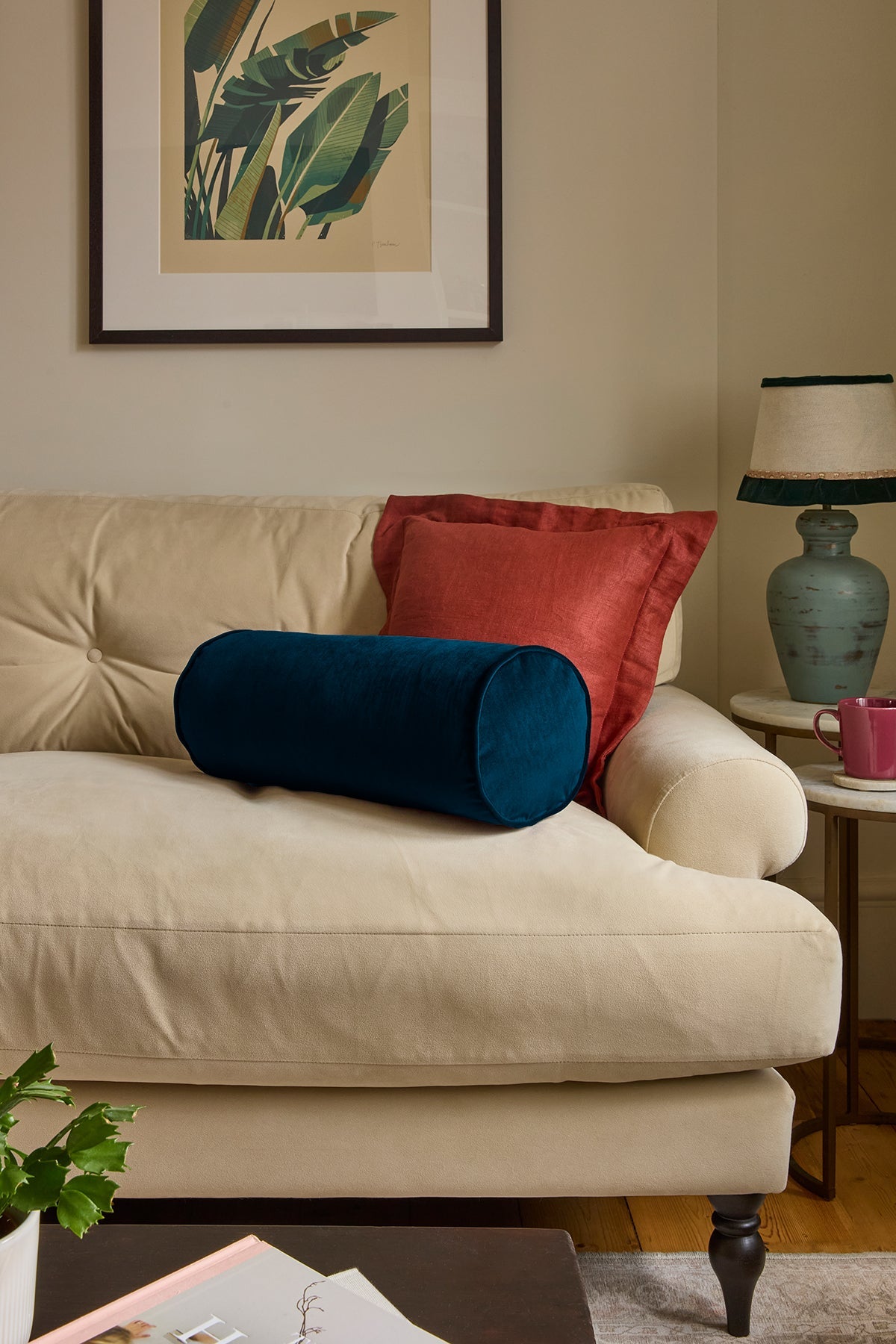 Beige sofa with blue cylindrical pillow and red cushion, framed artwork on wall