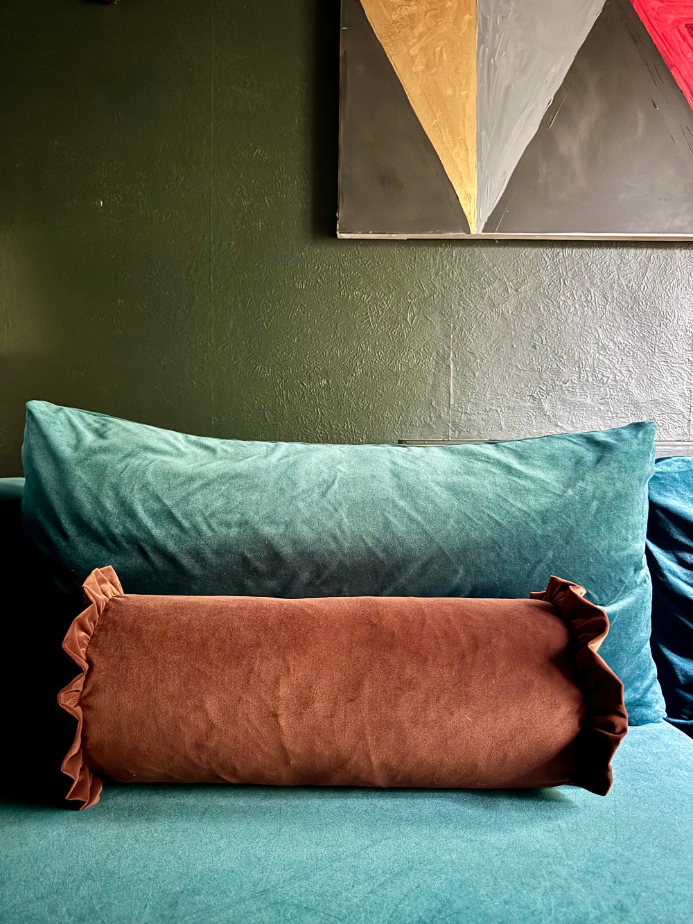 A brown ruffled lumbar pillow resting on a teal sofa in front of a larger teal pillow, set against a dark green wall with abstract geometric wall art.