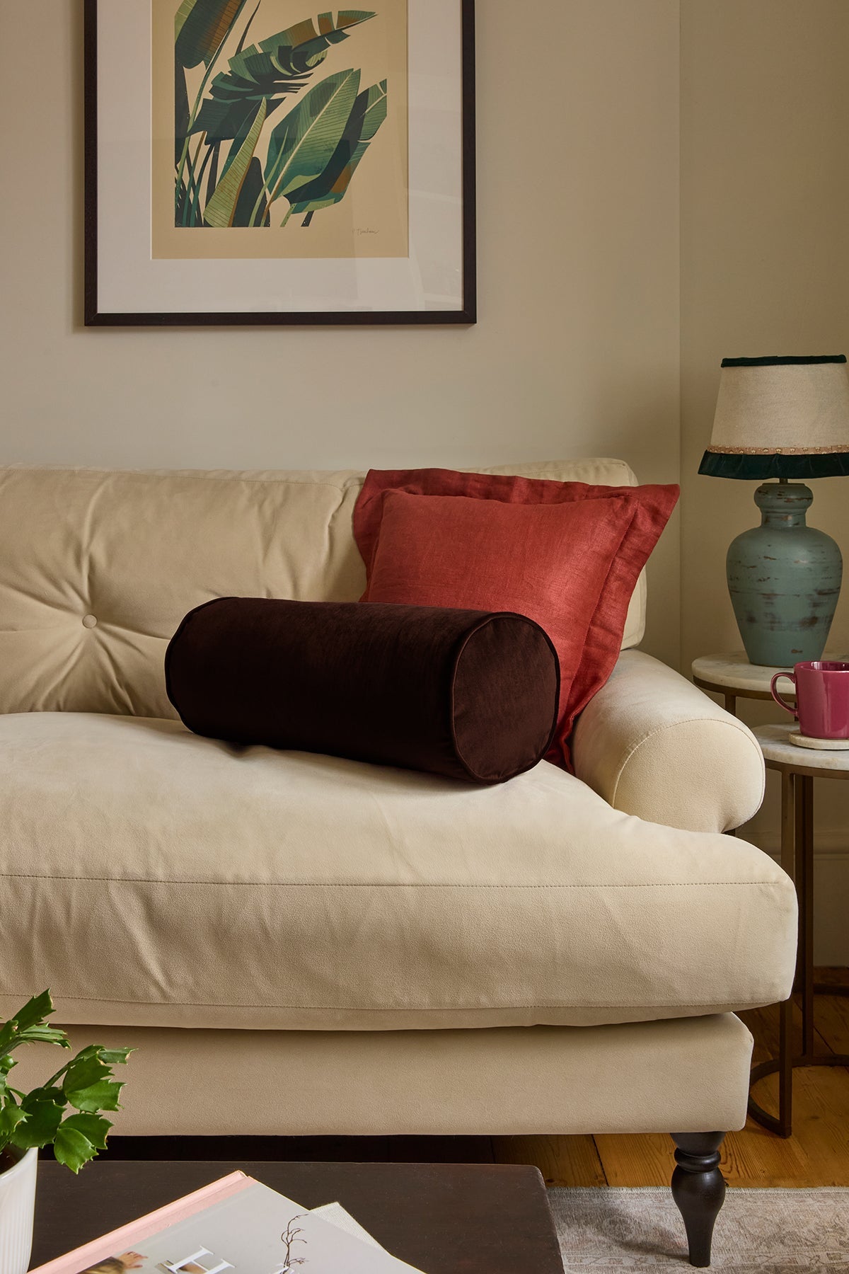 A cream-colored tufted sofa in a living room setting, styled with a dark brown cylindrical lumbar pillow, a rust-red square throw pillow, and a framed botanical print on the wall above.