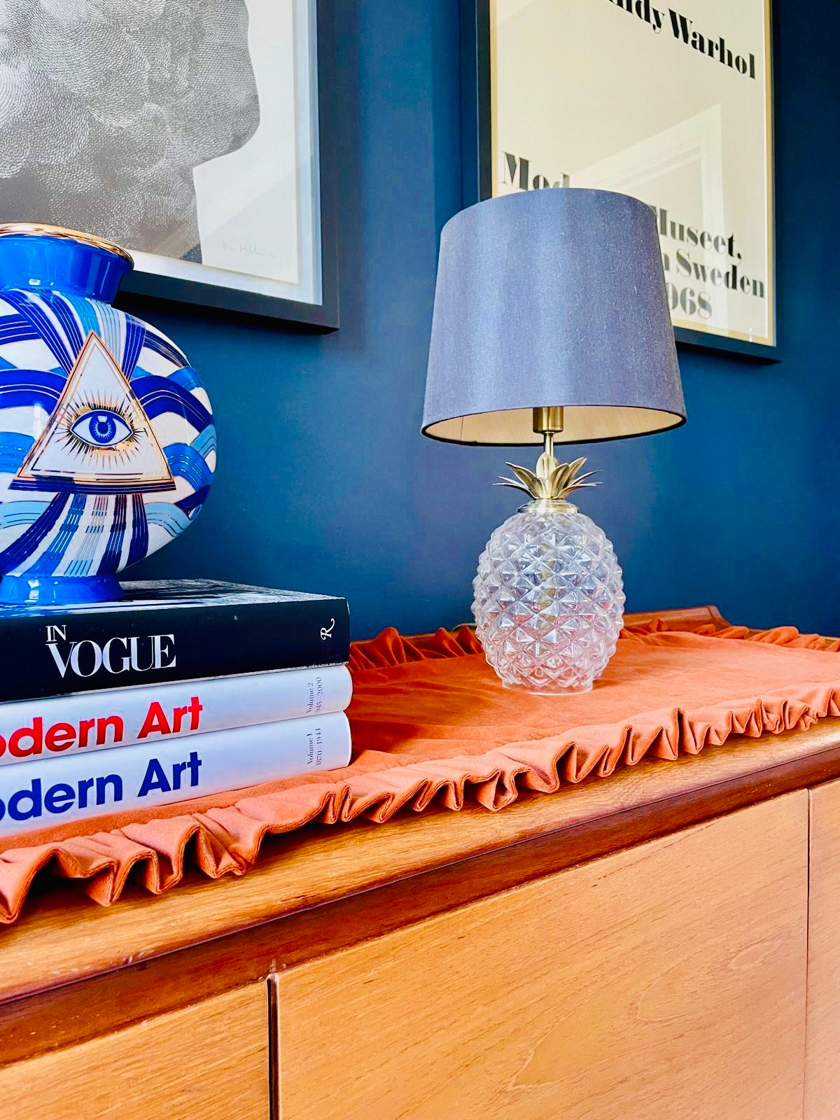 A pineapple-shaped clear glass table lamp with a gray fabric drum shade sits on a rust-orange ruffled fabric runner on a wooden sideboard. The scene is styled with books, a blue patterned vase, and framed wall art against a deep blue wall.
