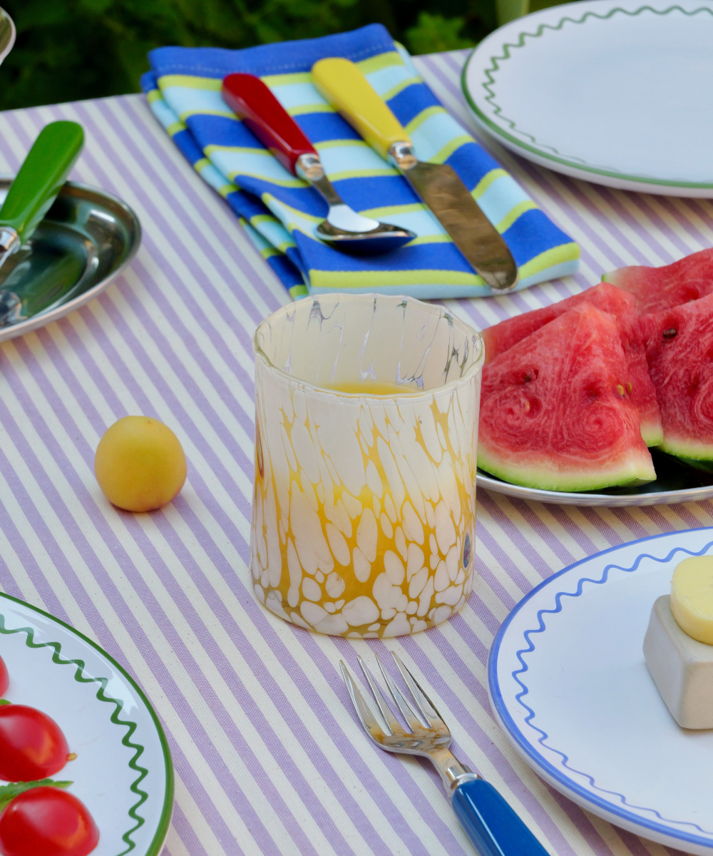 Outdoor table setting with watermelon, apricots, Yellow Murano glass tumbler and plates on a striped tablecloth.