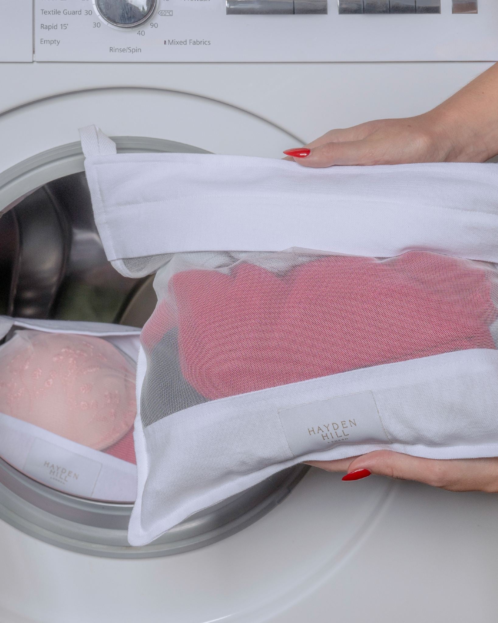 A close-up of a white mesh laundry bag being held open by hands with red nail polish, containing a pink knit garment, positioned in front of a white washing machine door.
