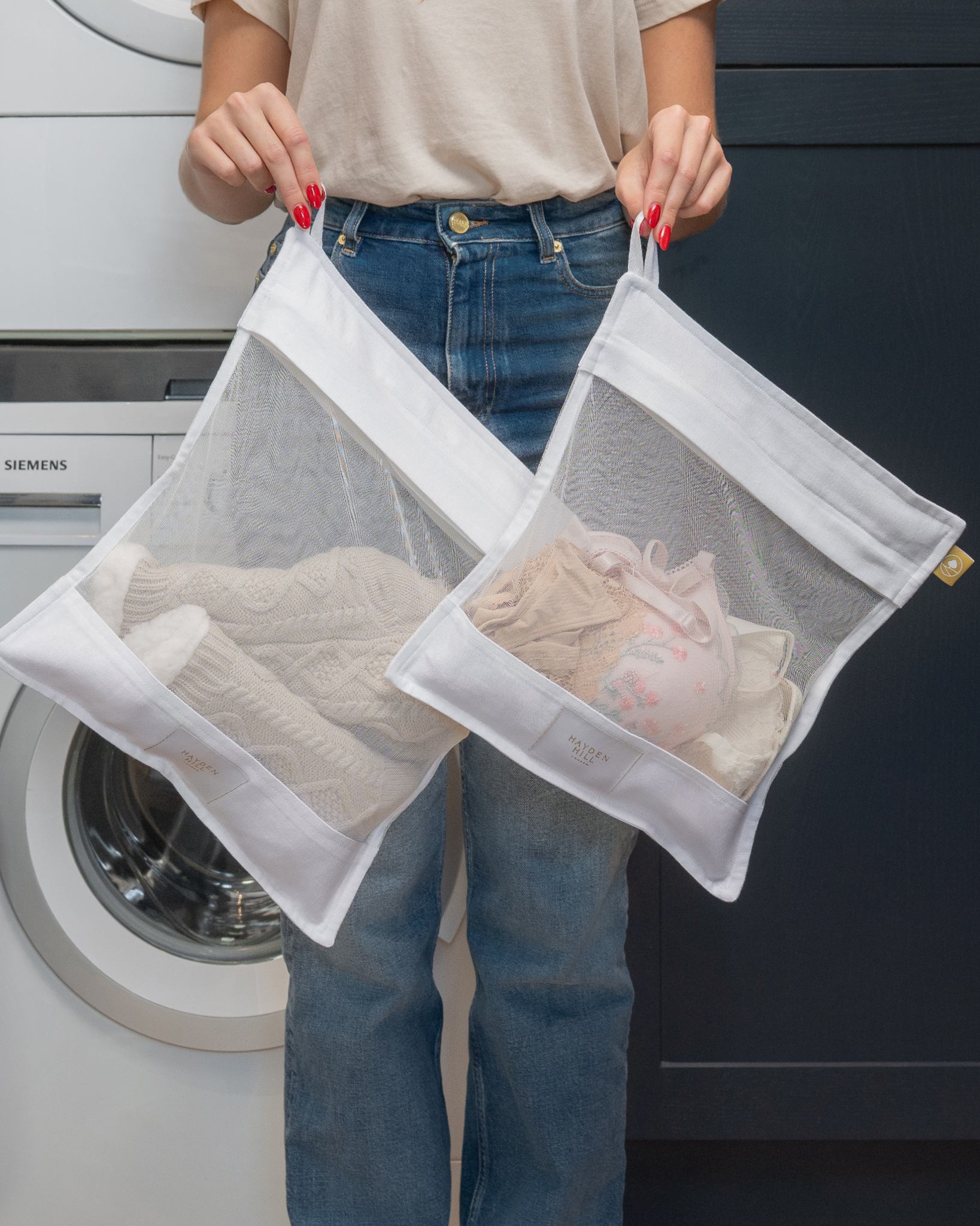 A person holding two white mesh laundry bags filled with folded clothes, standing in front of a washing machine and dark cabinet.
