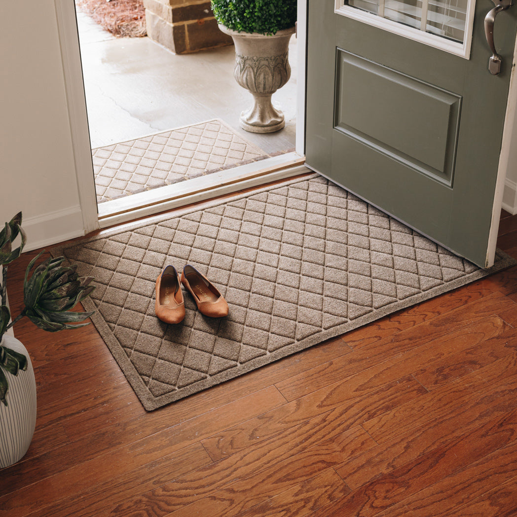 Camel WaterHog Argyle doormat positioned inside an open front door on a wooden floor with shoes nearby.