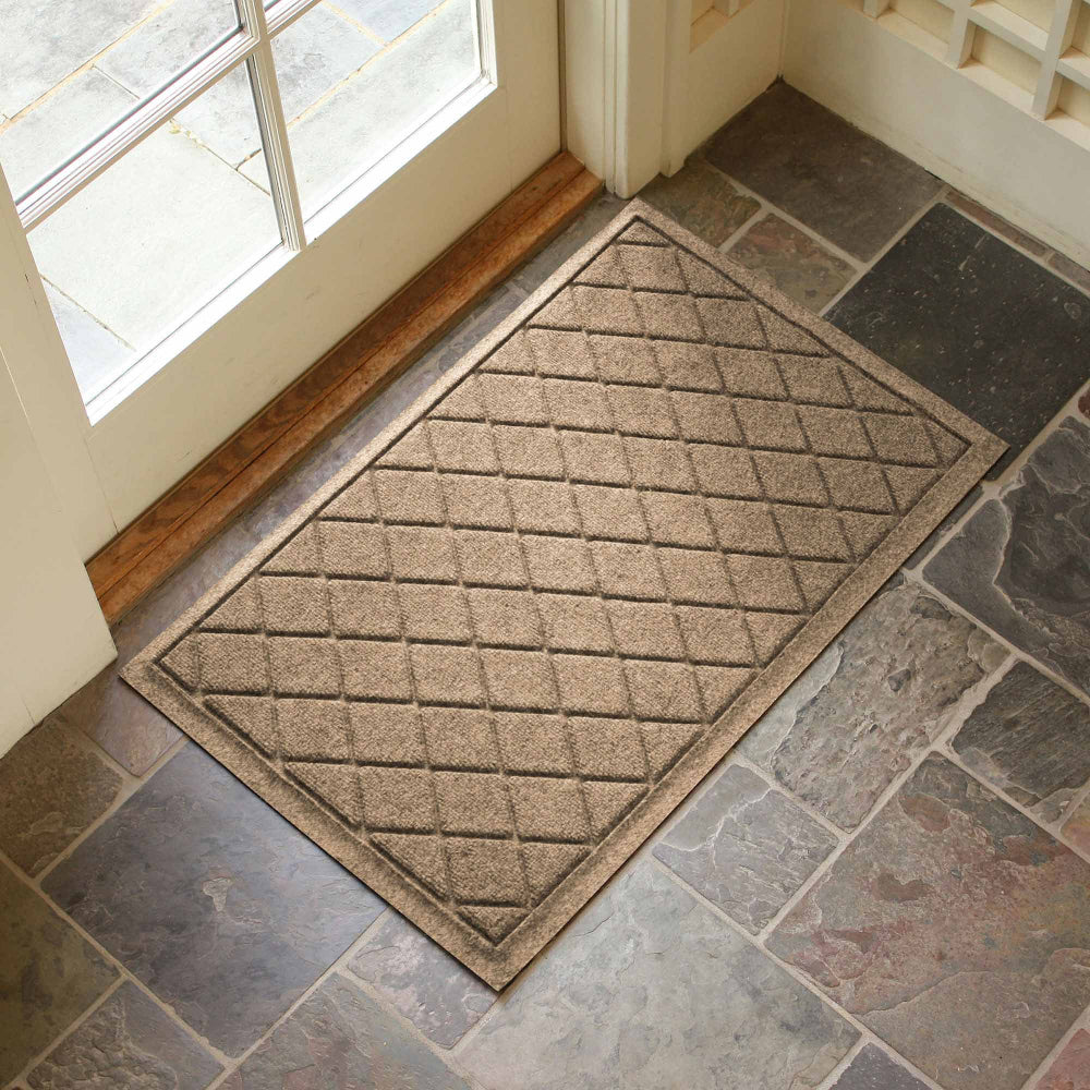 Camel WaterHog Argyle doormat positioned on a stone tiled floor beside a glass entrance door.