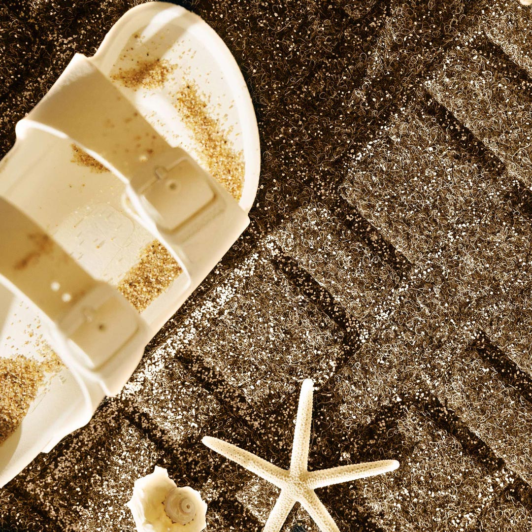 Close-up of sand on a textured WaterHog mat beside a light-coloured sandal and seashells.
