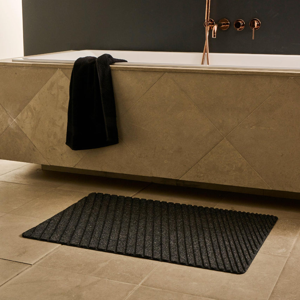 Charcoal WaterHog Gradient mat on a pale tiled bathroom floor beside a stone bathtub with a black towel.