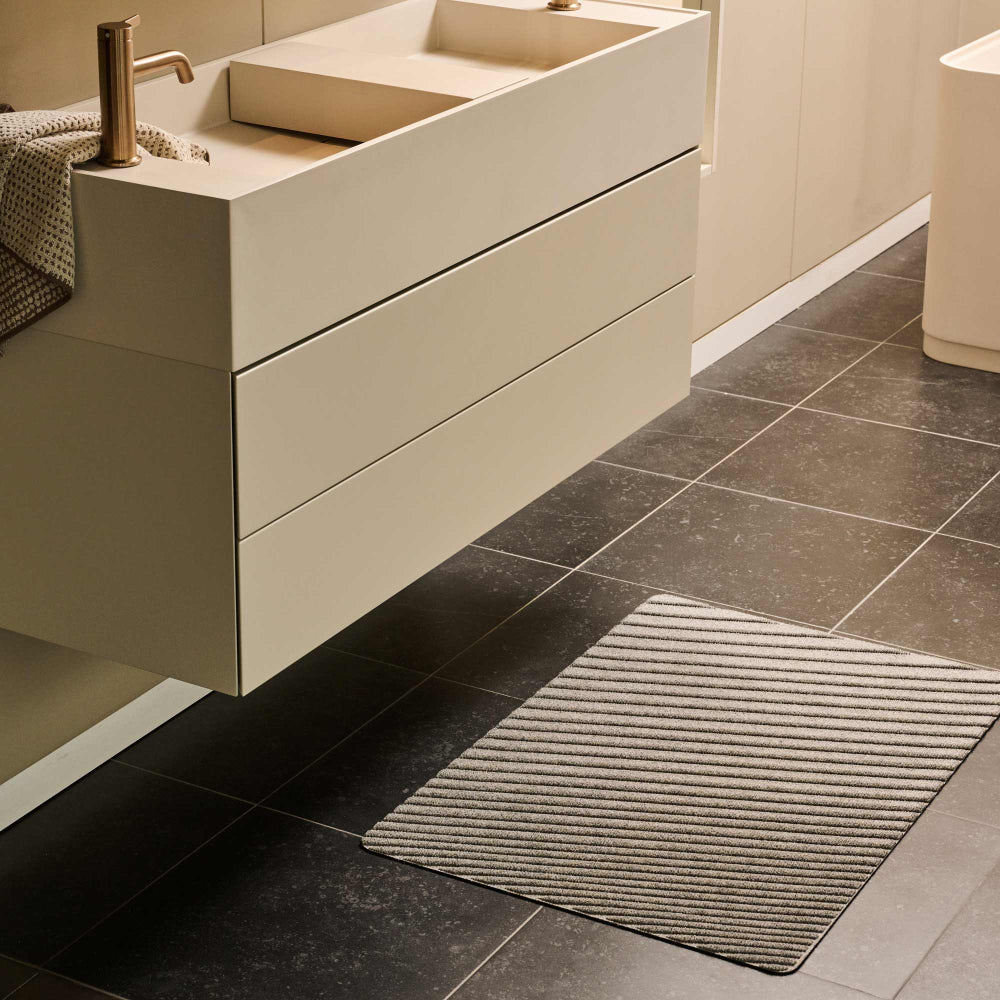 Grey WaterHog Gradient mat on dark tiled flooring beside a contemporary white bathroom vanity and basin.