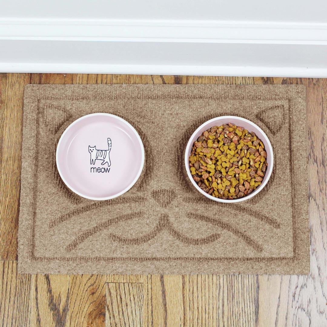 Overhead view of a Camel WaterHog Kitty Face Bowl Mat with two pink ceramic bowls, one filled with dry cat food.