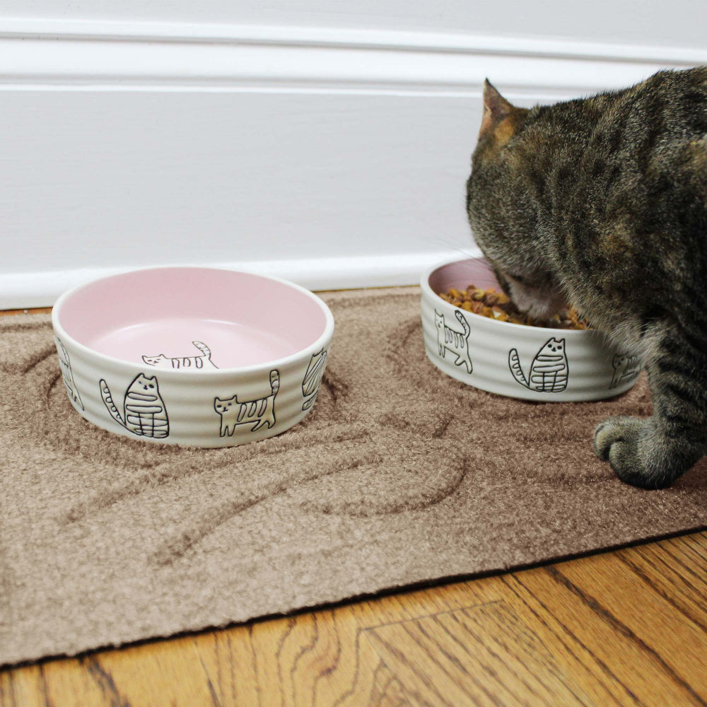 Tabby cat eating dry food from a ceramic bowl on a Camel WaterHog feeding mat beside a matching empty bowl.