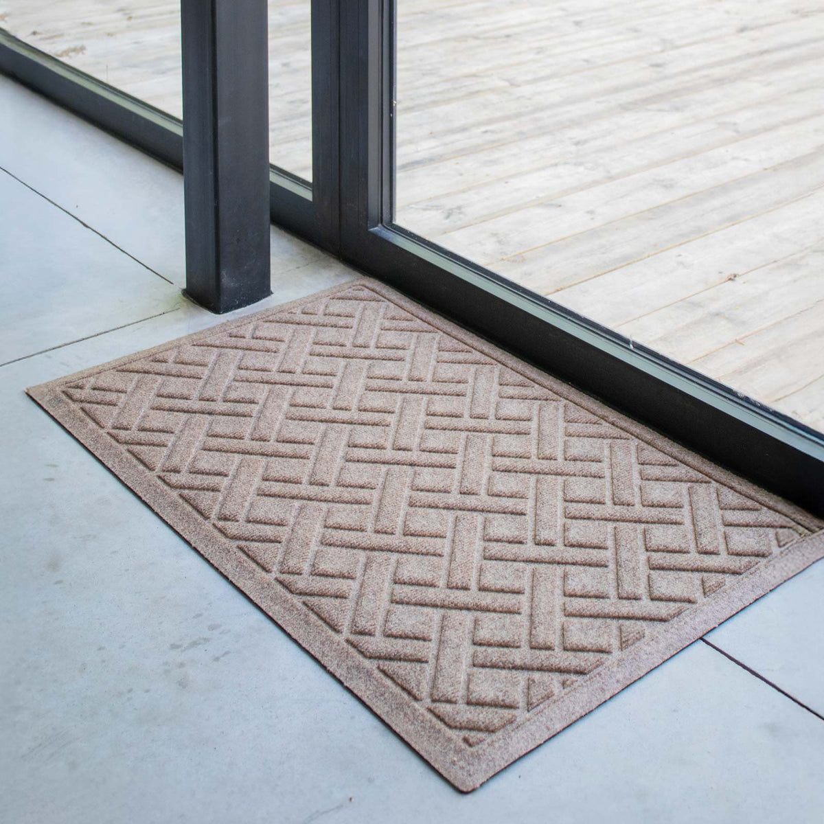 Camel WaterHog Lattice doormat positioned on a pale floor next to a black-framed glass patio door.