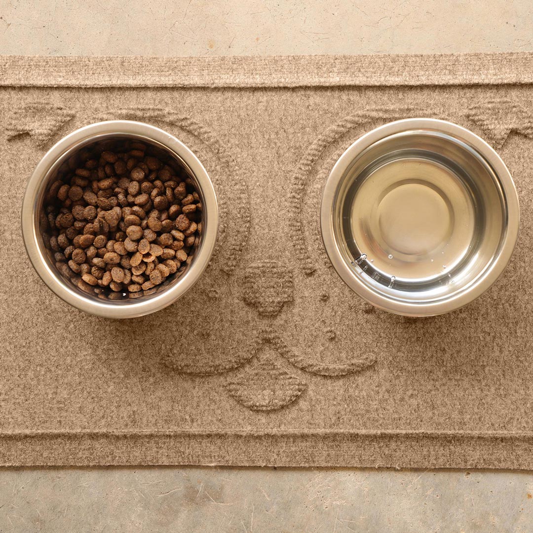 Overhead view of a Camel WaterHog Puppy Face feeding mat with two stainless steel bowls containing dry dog food and water.