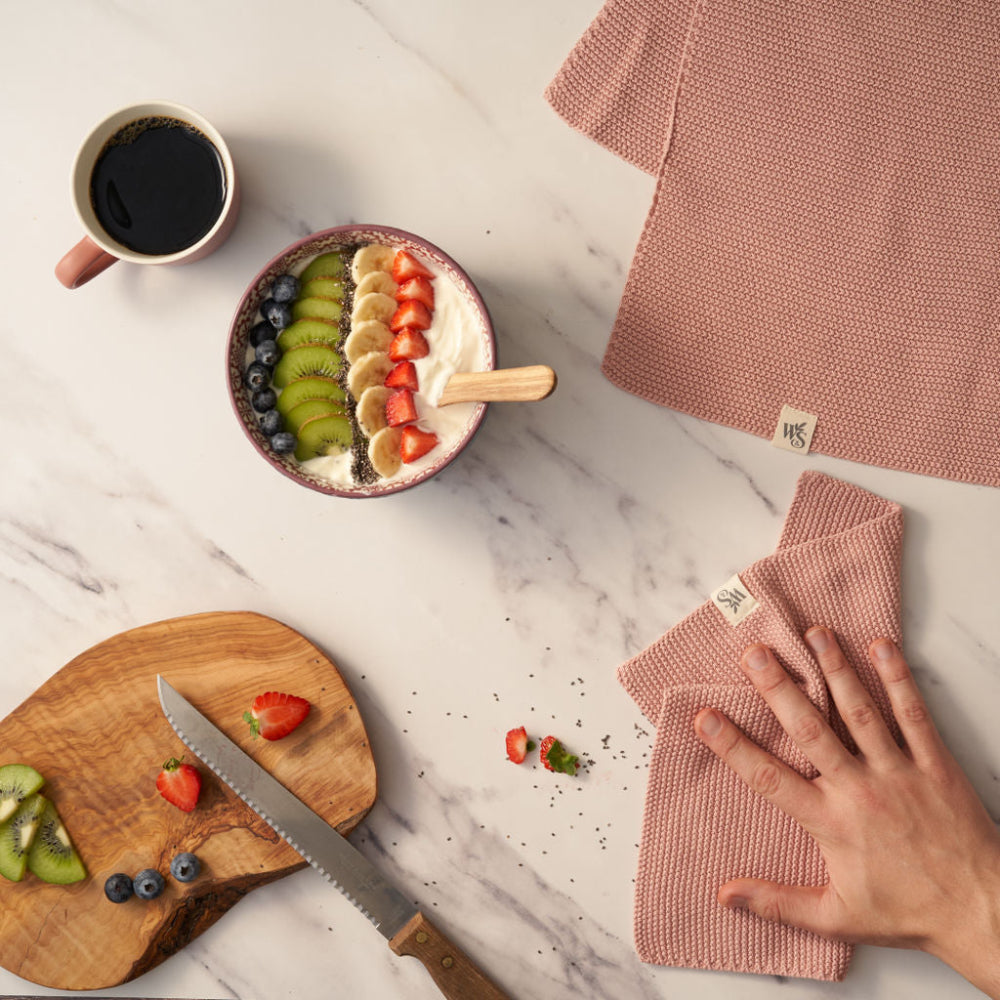 Fruit bowl with a cup of coffee, wooden cutting board, and pink dishcloths on a marble surface.