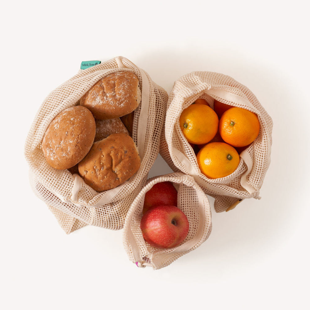 Three reusable mesh bags with bread, apples, and oranges on a white background