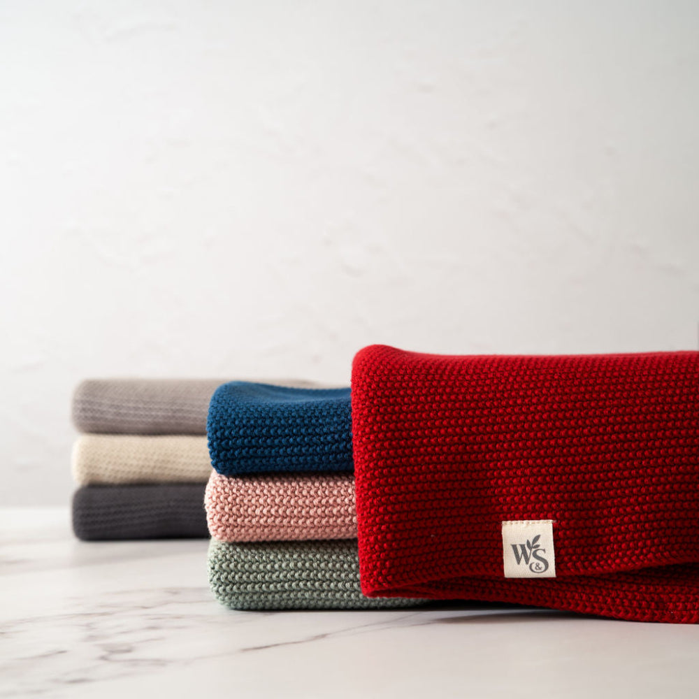 Stack of folded organic hand towels in red, blue, pink, and grey on a light surface with a white background.