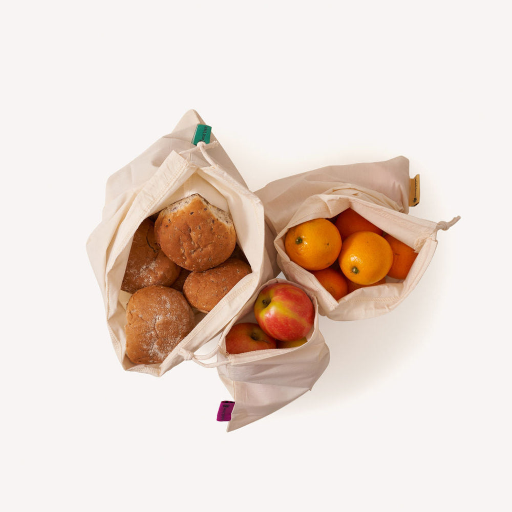 Three reusable organic cotton bags filled with bread and fruits on a white background