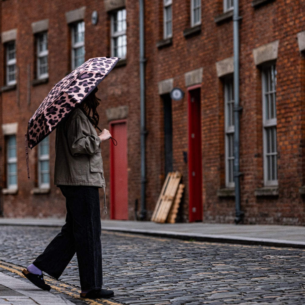 Person walking on a cobblestone street with a leopard print umbrella in front of brick buildings.