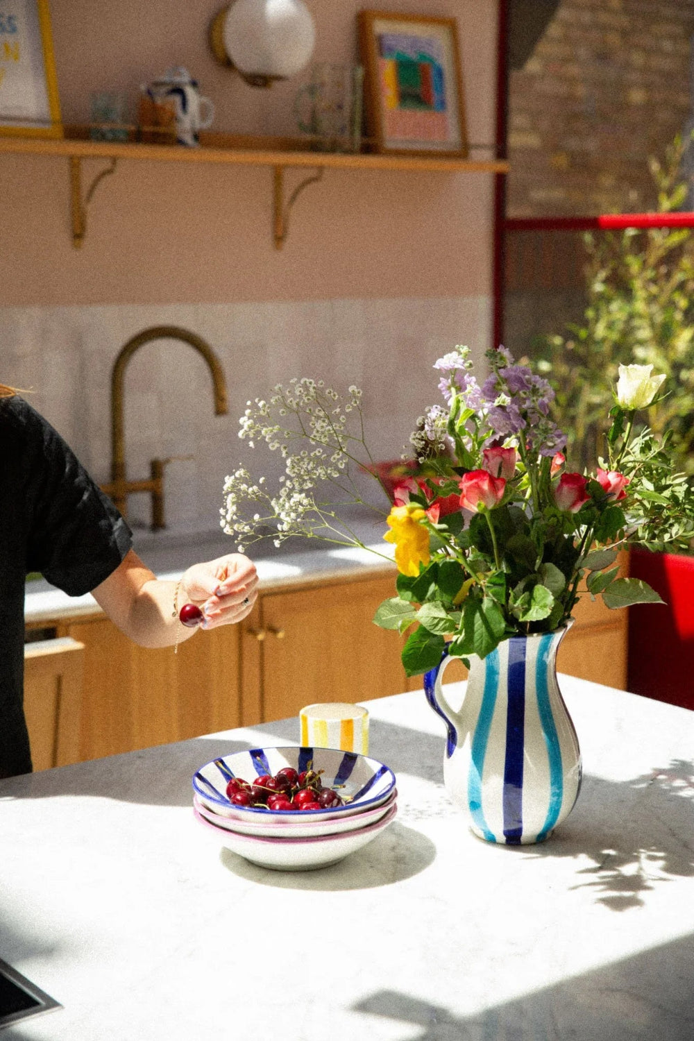 A kitchen scene with a person's arm visible on the left, holding a small white flower sprig beside a striped ceramic pitcher filled with colorful flowers and a matching striped bowl of red cherries on a light countertop.