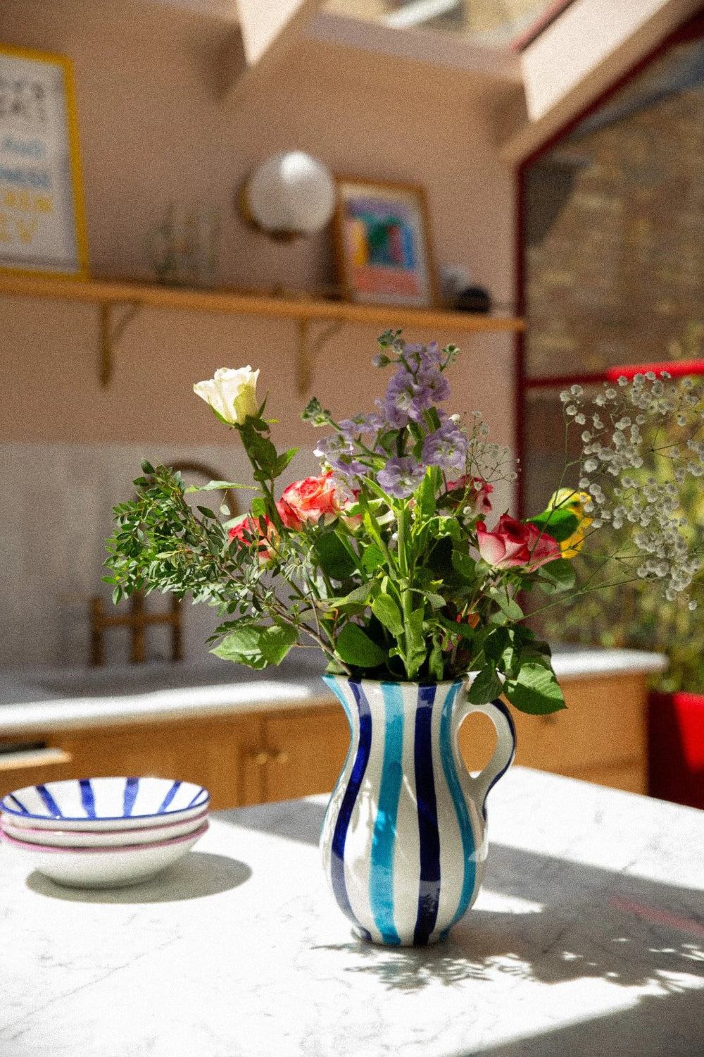 A ceramic pitcher vase with vertical blue and white stripes, filled with a bouquet of fresh wildflowers, set on a light marble countertop in a warm, sunlit kitchen.