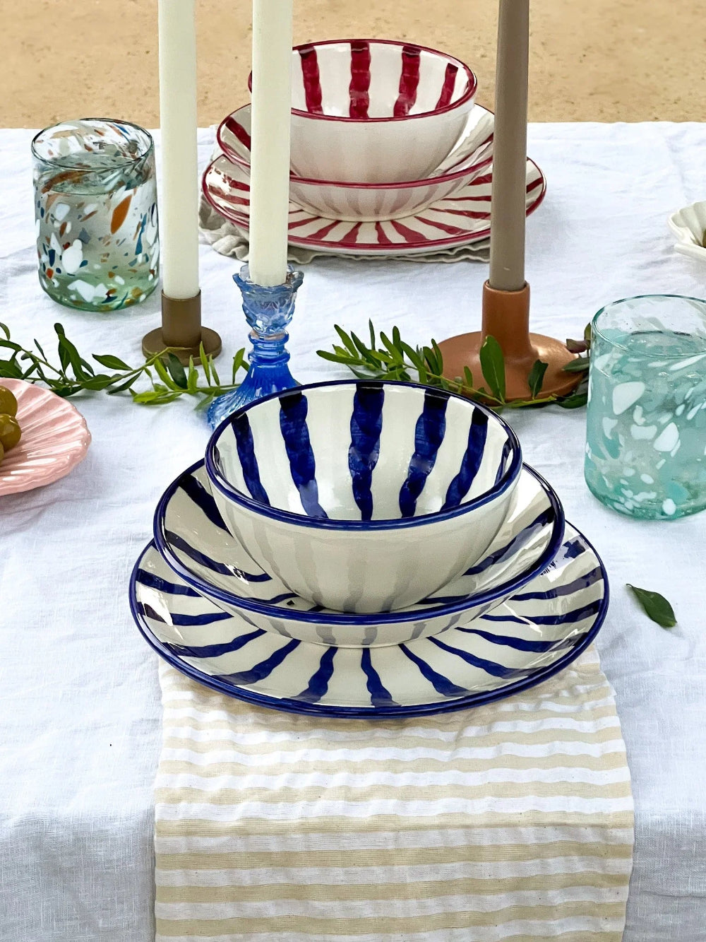 A styled table setting featuring a stack of white ceramic bowls and matching plates with bold blue and red stripes, set on a white tablecloth with candles, glasses, and greenery.