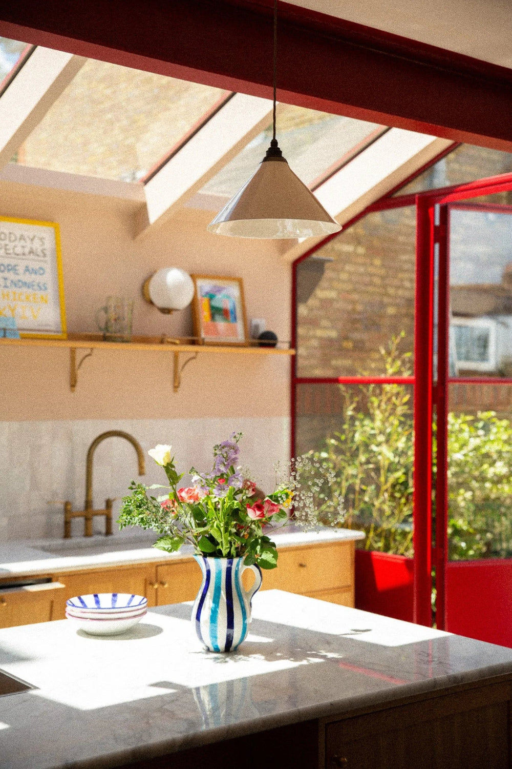 A bright kitchen interior with a marble countertop in the foreground, a blue-and-white striped vase holding colorful flowers, wooden cabinetry, a sink, and a red-framed glass door opening to greenery outside.