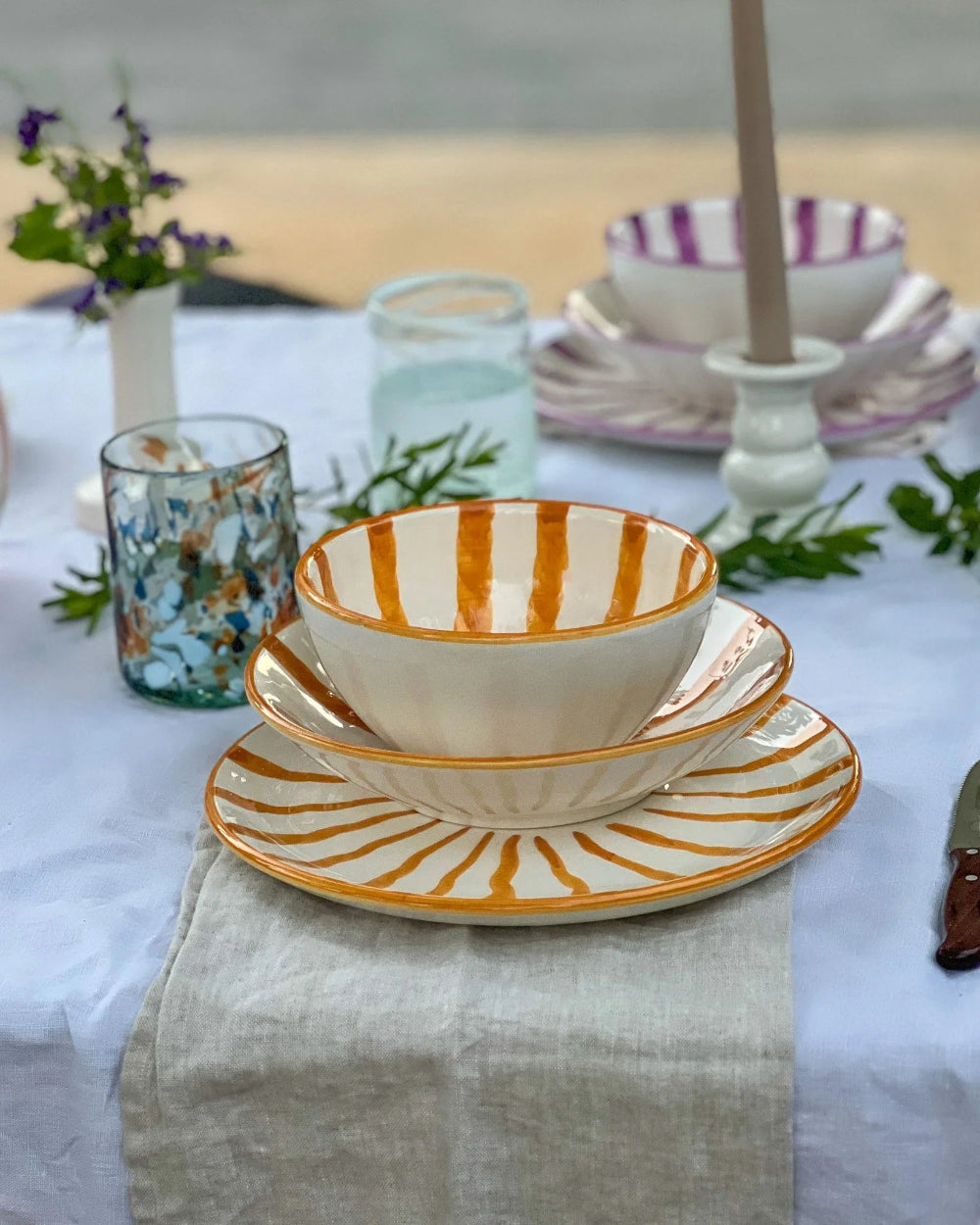 A set of white ceramic tableware with orange stripes and sunburst patterns, styled on a white tablecloth with a linen runner, glassware, and greenery.